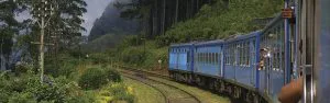 Blauwe trein rijdt door een groene, bergachtige omgeving met dichte bossen en mistige heuvels op de achtergrond.