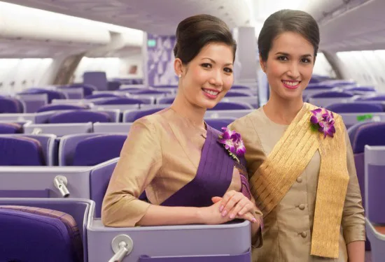 Twee stewardessen van Thai Airways in traditionele, elegante uniformen met bloemen op hun schouder, staan glimlachend in een luxe vliegtuigcabine met paarse stoelen.