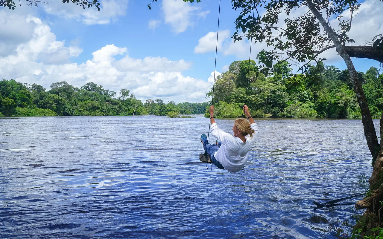 Reiziger slingert aan een touw over een brede rivier, omringd door dichtbegroeid regenwoud en een blauwe lucht.