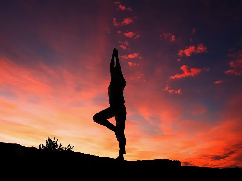 Een silhouet van een persoon in een yogahouding op één been, tegen een kleurrijke zonsondergang met oranje en paarse wolken.