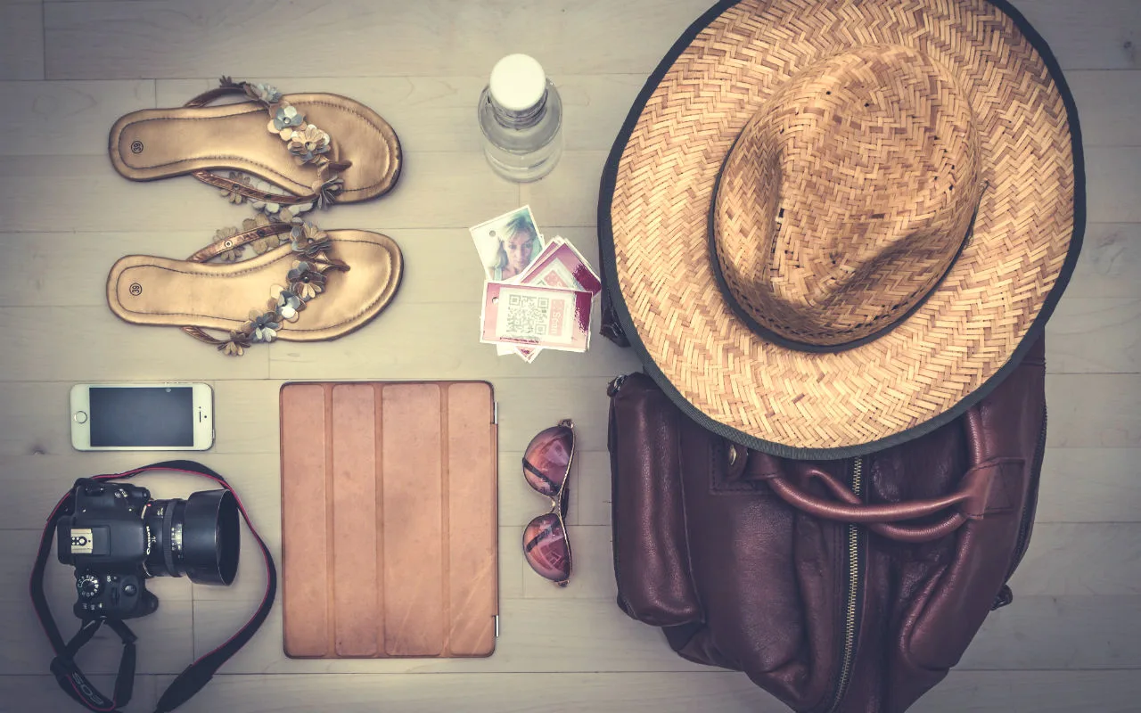 Vakantiebenodigdheden op een houten vloer: sandalen, waterfles, geld, zonnebril, rieten hoed, leren rugzak, tablet, smartphone en fotocamera.