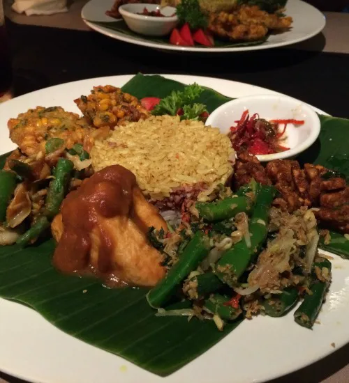 Traditioneel Indonesisch gerecht op een wit bord met bananenblad, bestaande uit gele rijst, groentegerechten, tofu met pindasaus, tempeh en verschillende bijgerechten.