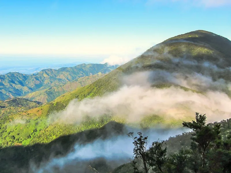Uitzicht op een berglandschap met groene hellingen en laaghangende wolken, met in de verte de blauwe lucht en zee.