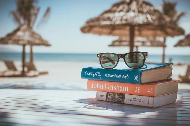 Twee boeken met een zonnebril erop liggen op een tafel aan het strand, met rieten parasols en zee op de achtergrond.