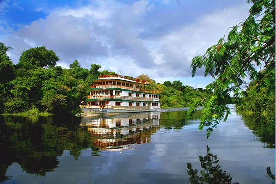 Wit riviercruiseschip met meerdere dekken vaart door een tropische rivier omringd door jungle.