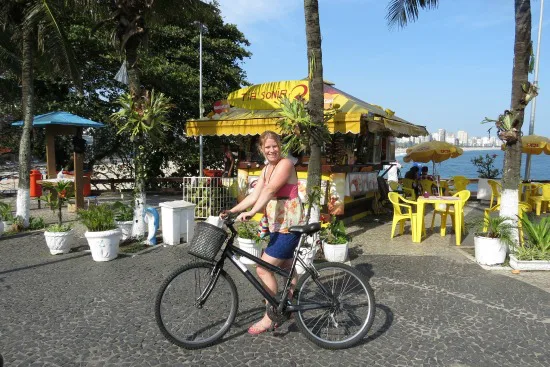 Vrouw fietst langs een kleurrijk strandtentje aan de boulevard, met uitzicht op zee en palmbomen op de achtergrond