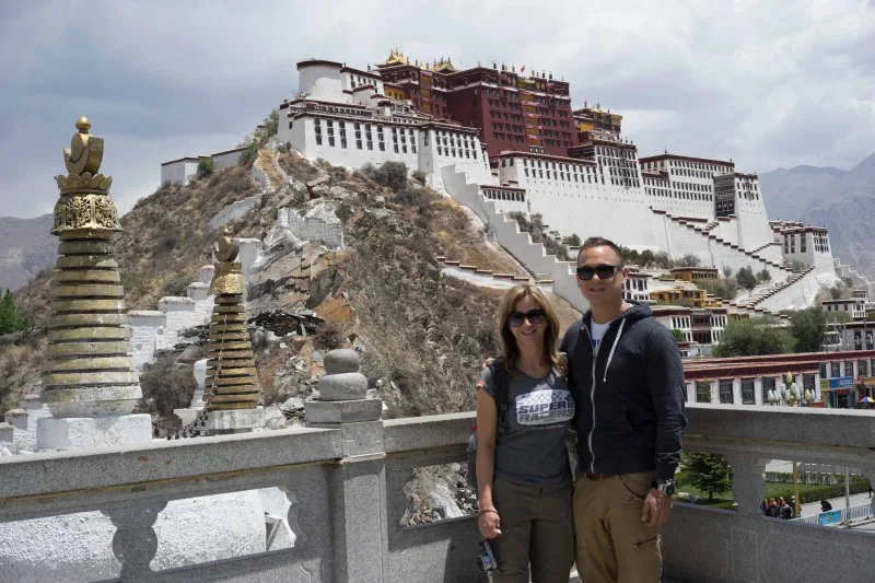 Man en vrouw poseren voor de Potala-paleis in Lhasa, Tibet, met witte muren en rood-gouden daken op een heuvel.
