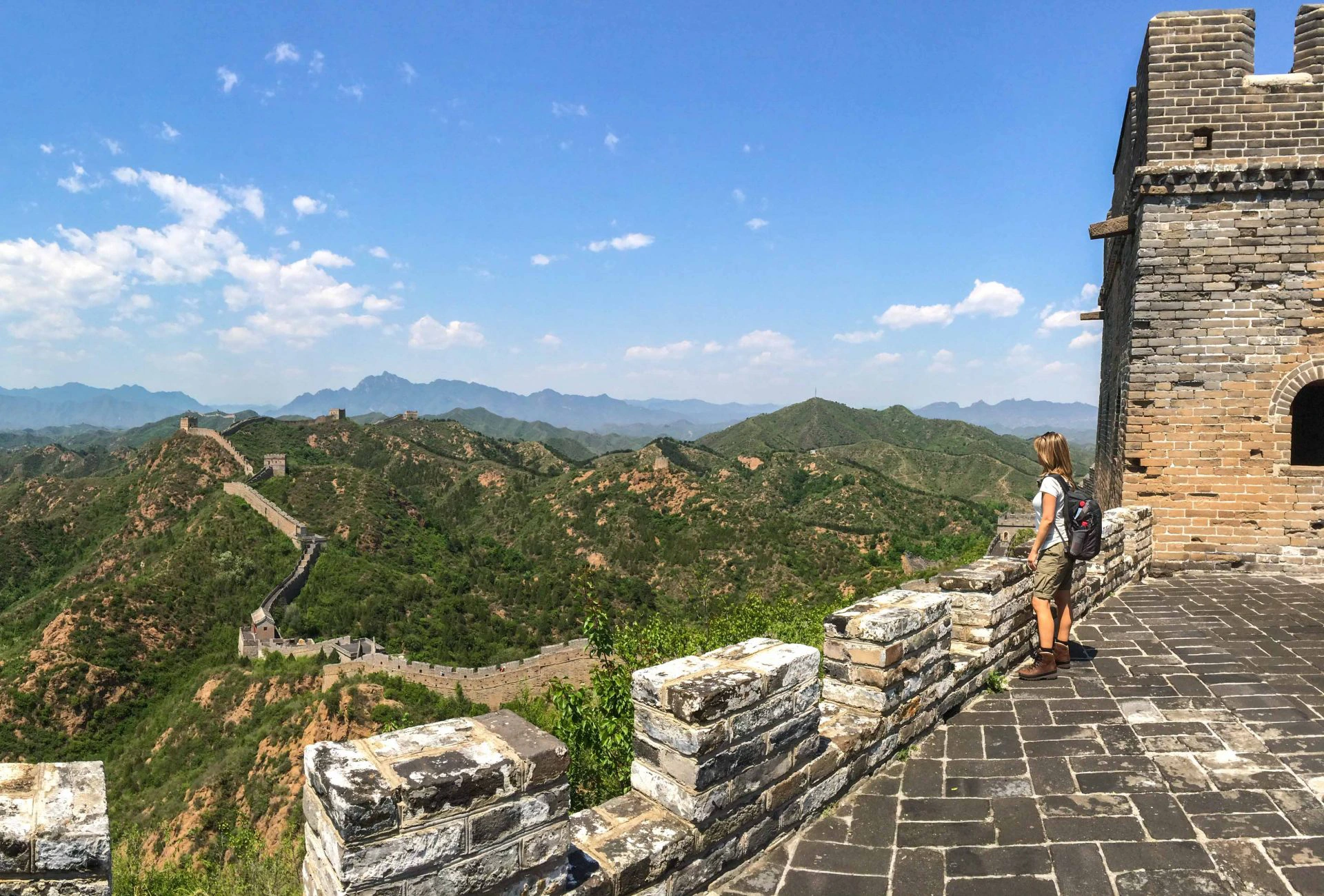Reiziger met rugzak kijkt uit over de kronkelende Chinese Muur en groene berglandschappen onder een blauwe lucht met witte wolken