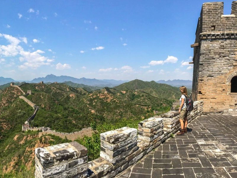 Reiziger met rugzak kijkt uit over de kronkelende Chinese Muur en groene berglandschappen onder een blauwe lucht met witte wolken