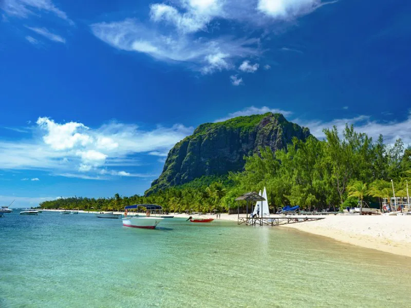 Witte zandstrand met helderblauw water en kleine bootjes, omringd door palmbomen en met de imposante Le Morne Brabant berg op de achtergrond in Mauritius.