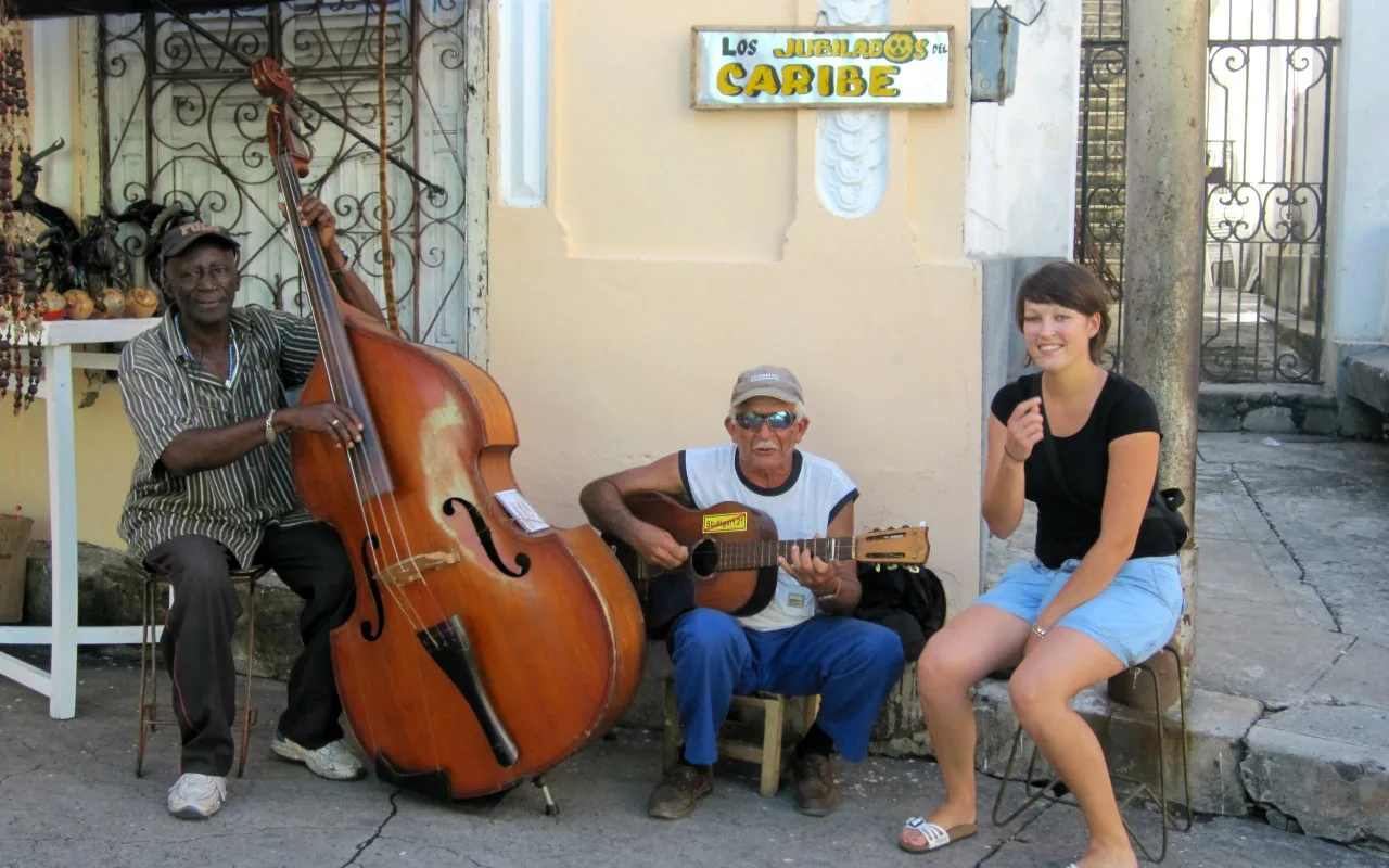 Twee straatmuzikanten spelen contrabas en gitaar, terwijl een vrouw naast hen zit en lacht, met op de achtergrond een bord met de tekst "Los Naranjos Caribe".