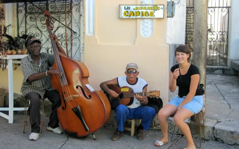 Twee straatmuzikanten spelen contrabas en gitaar, terwijl een vrouw naast hen zit en lacht, met op de achtergrond een bord met de tekst "Los Naranjos Caribe".