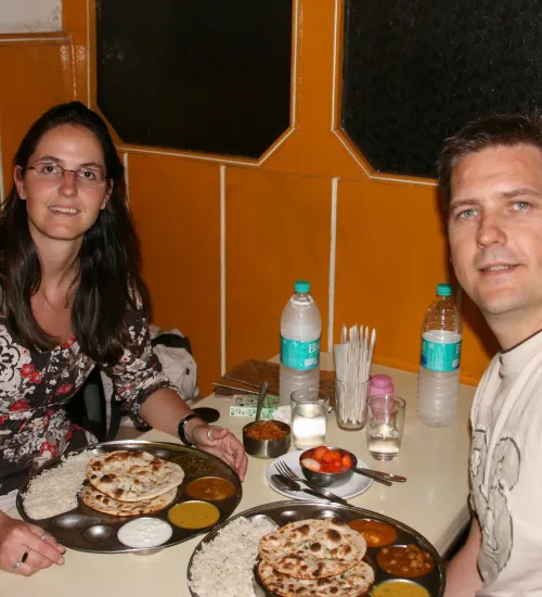 Twee mensen zitten in een eenvoudig restaurant in India met voor hen een traditioneel Indiaas gerecht op een metalen bord, bestaande uit naanbrood, rijst en verschillende curry’s.