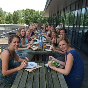 Grote groep mensen zit aan lange houten picknicktafels op een balkon, samen etend en lachend in de richting van de camera.