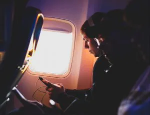 Twee vliegtuigpassagiers zitten naast elkaar, één kijkt naar zijn telefoon bij het raam met zonlicht dat naar binnen schijnt