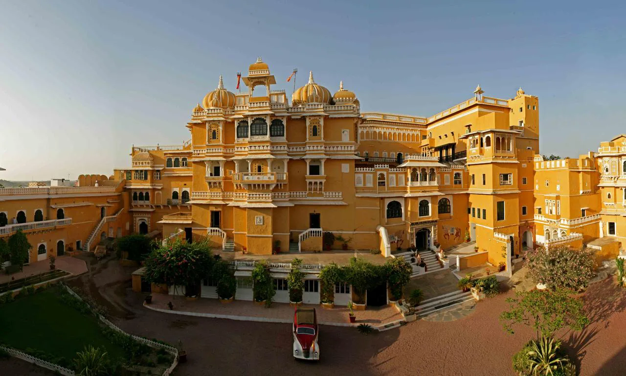 Een groot geel paleis in Rajasthan, India, met meerdere koepels, bogen en balkons, omringd door een binnenplaats met groen en een rode riksja.