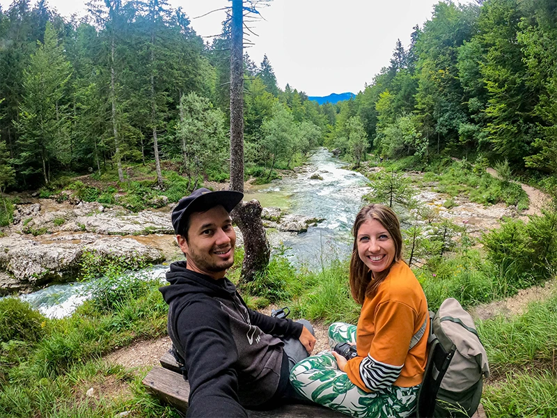 Twee reizigers zitten op een bankje in een bosrijke omgeving langs een rivier, lachend in de camera met groene bomen en stromend water op de achtergrond.