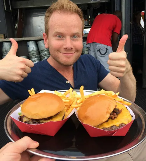 Man glimlacht naar de camera terwijl hij twee hamburgers met friet op een dienblad vasthoudt en beide duimen omhoog steekt.