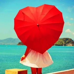 Vrouw in een witte jurk staat aan zee met een groot rood hartvormig paraplu, terwijl op de achtergrond helderblauw water, groene heuvels en een zonnige lucht te zien zijn.