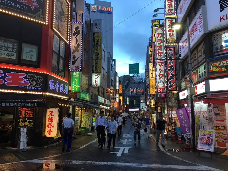 Een levendige straat in Japan bij schemering, vol felverlichte neonreclames en Japanse tekens, met mensen die langs ramen van restaurants en winkels slenteren.
