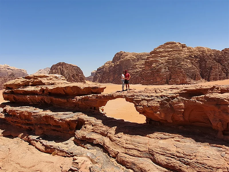 Twee mensen staan op een natuurlijke rotsbrug in de woestijn van Wadi Rum in Jordanië, met op de achtergrond hoge roodbruine rotsformaties onder een strakblauwe lucht.