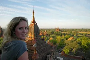 Vrouw kijkt over haar schouder vanaf een tempel in Bagan, Myanmar, met uitzicht op eeuwenoude pagodes en groene vlaktes in de avondzon.