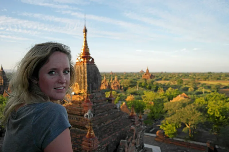 Vrouw kijkt over haar schouder vanaf een tempel in Bagan, Myanmar, met uitzicht op eeuwenoude pagodes en groene vlaktes in de avondzon.