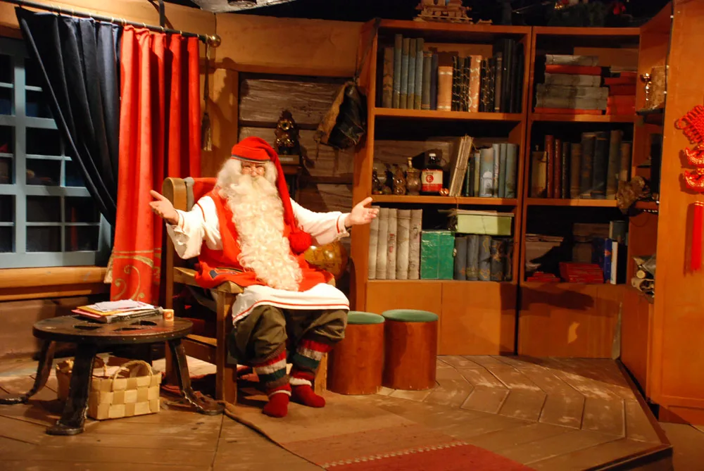 Een man verkleed als kerstman met lange witte baard zit in een gezellige kamer vol boeken en houten meubels in Rovaniemi, Lapland.