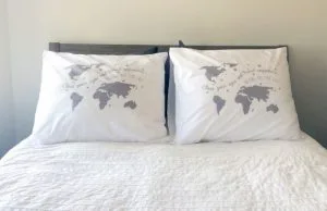 Twee witte kussens op een bed, elk bedrukt met een wereldkaart en de tekst “Close your eyes and travel anywhere”, tegen een lichtgrijze muur.