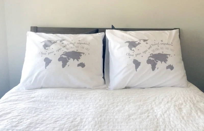 Twee witte kussens op een bed, elk bedrukt met een wereldkaart en de tekst “Close your eyes and travel anywhere”, tegen een lichtgrijze muur.