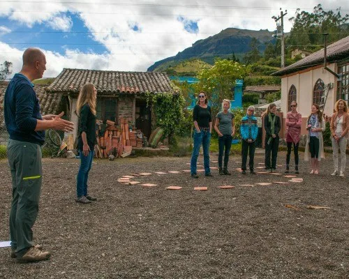 Groep mensen staat in een kring buiten op een erf in de bergen terwijl een man iets uitlegt.