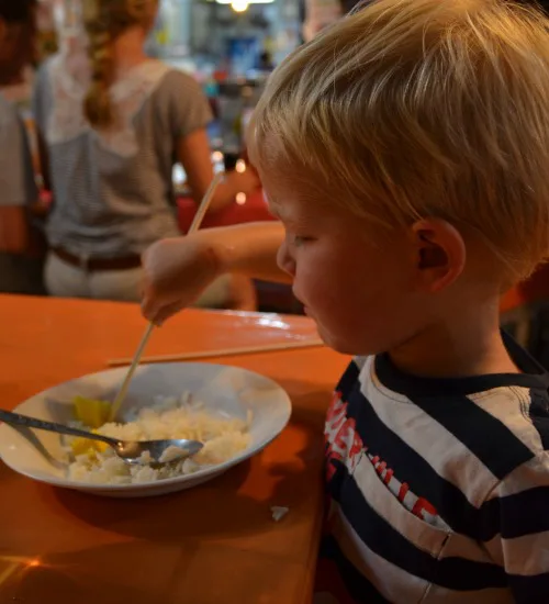 Jonge jongen eet rijst met stokjes aan een tafel op een drukke avondmarkt.