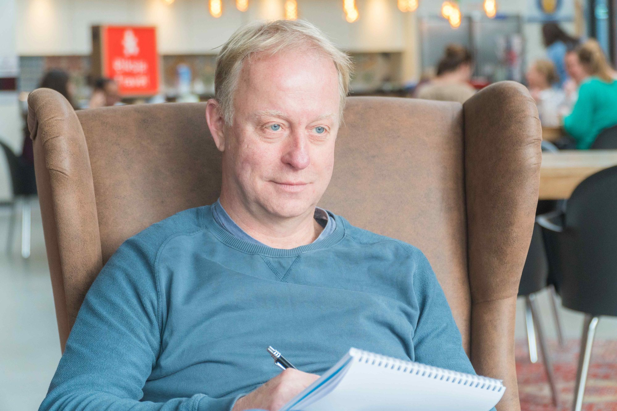 Man zit in een bruine stoel met een notitieboek op schoot en een pen in de hand, in een drukke ruimte met mensen op de achtergrond.