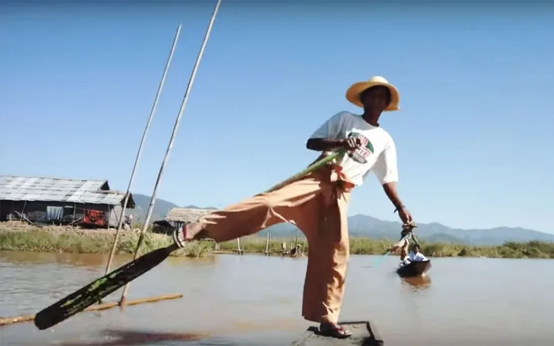 Man op een boot die met één been roeit op een meer, met op de achtergrond een houten huis op palen, groene oevers en bergen onder een heldere blauwe lucht.