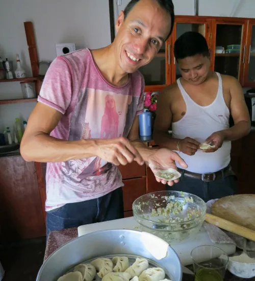 Twee mannen in een keuken die samen dumplings maken; de voorste man lacht naar de camera terwijl hij een dumpling vult, de andere man is geconcentreerd bezig in de achtergrond.