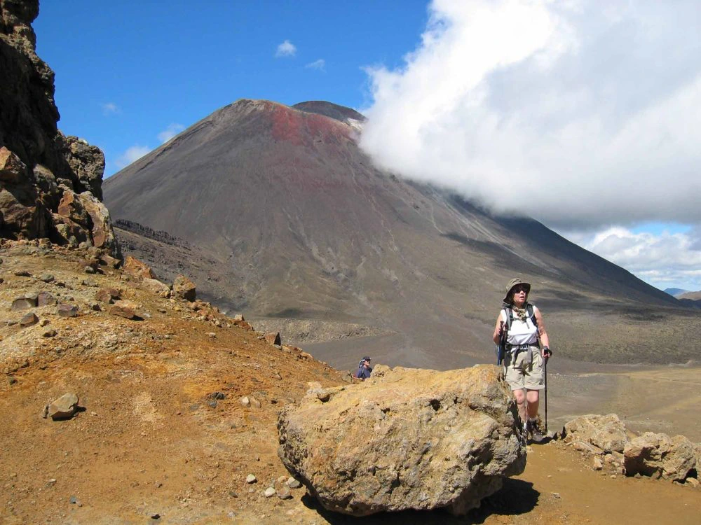Wandelaar met hoed en rugzak op een rotsachtig pad, met de donkere vulkaan Mount Ngauruhoe in Tongariro National Park, Nieuw-Zeeland, op de achtergrond.