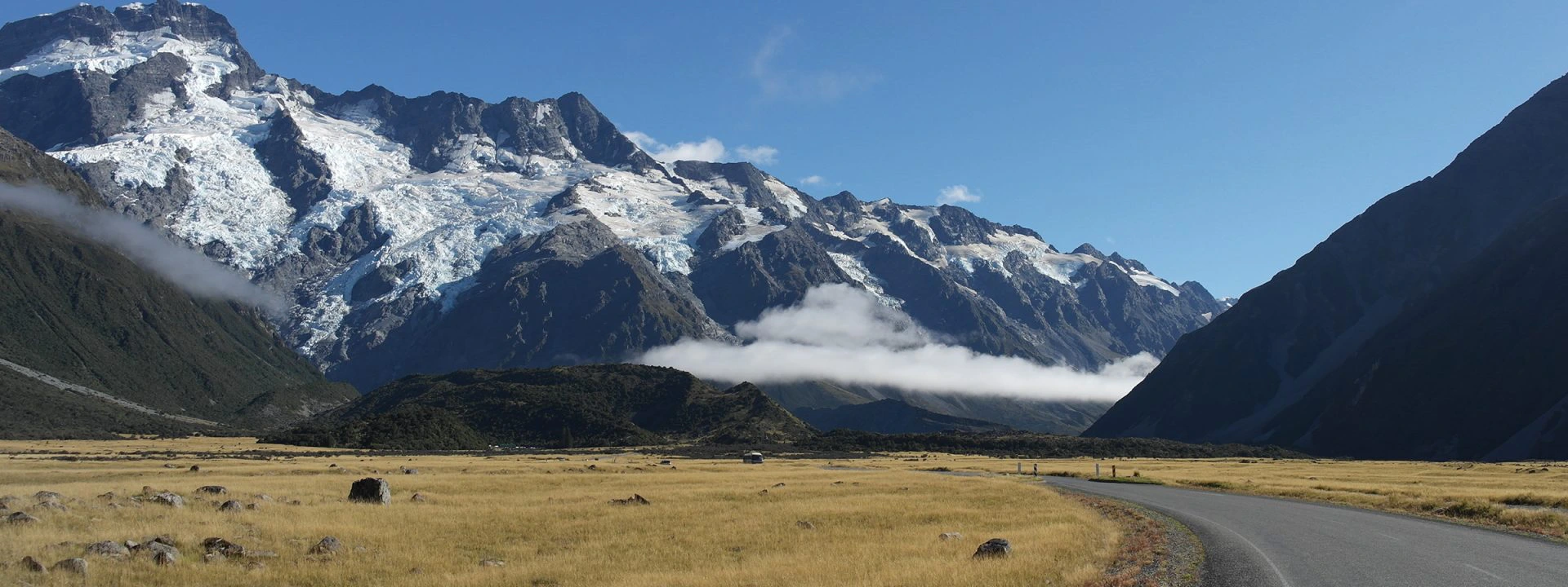 Berglandschap met besneeuwde toppen en gletsjers, een open vlakte met geel gras en een kronkelende weg op de voorgrond.