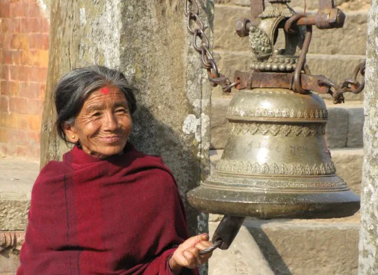 Oudere Nepalese vrouw in een rode sjaal glimlacht terwijl ze naast een grote bronzen tempelbel staat, met stenen trappen en muren op de achtergrond.