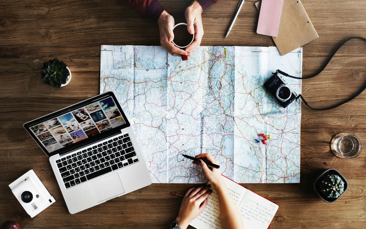 Tafel met een uitgevouwen landkaart, laptop, camera, notitieboek en handen die een reisroute plannen.