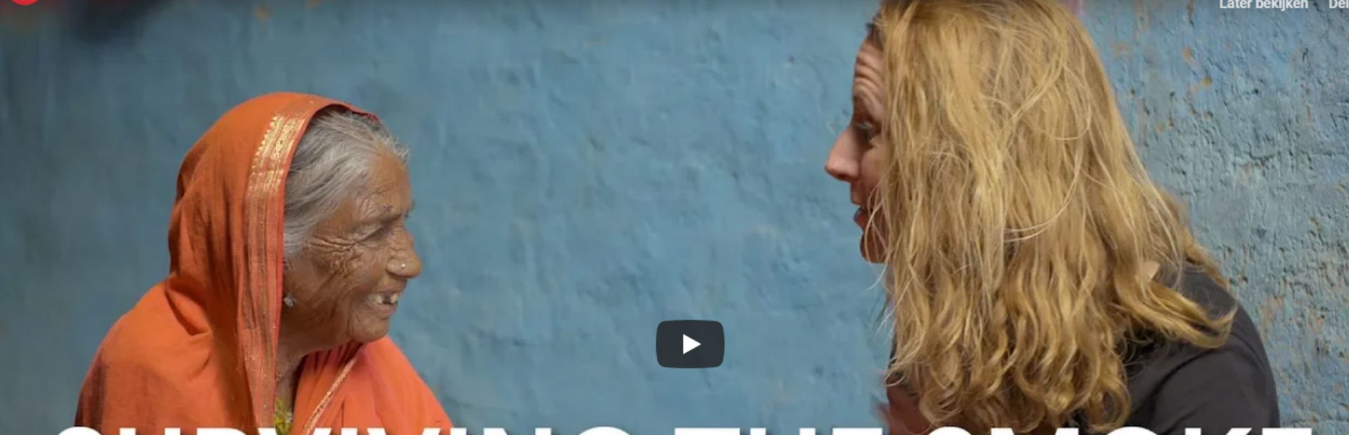 Oudere vrouw in oranje sari glimlacht terwijl ze met een blonde vrouw praat tegen een blauwe muur.