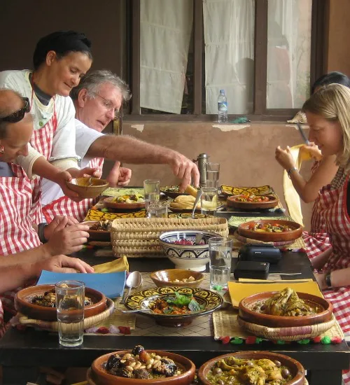 Groep reizigers en een lokale vrouw genieten samen van een maaltijd tijdens een kookworkshop, met kleurrijke gerechten op tafel en iedereen in rood-wit gestreepte schorten.