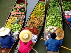 Vier houten boten op een drijvende markt, gevuld met tropisch fruit zoals watermeloen, bananen en groene appels, met verkopers in kleurrijke kleding en rieten hoeden.