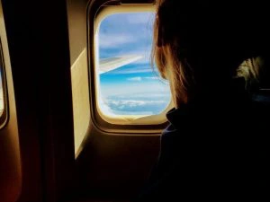 Vrouw kijkt uit het raam van een vliegtuig naar een blauwe lucht met wolken en de vleugel van het toestel.