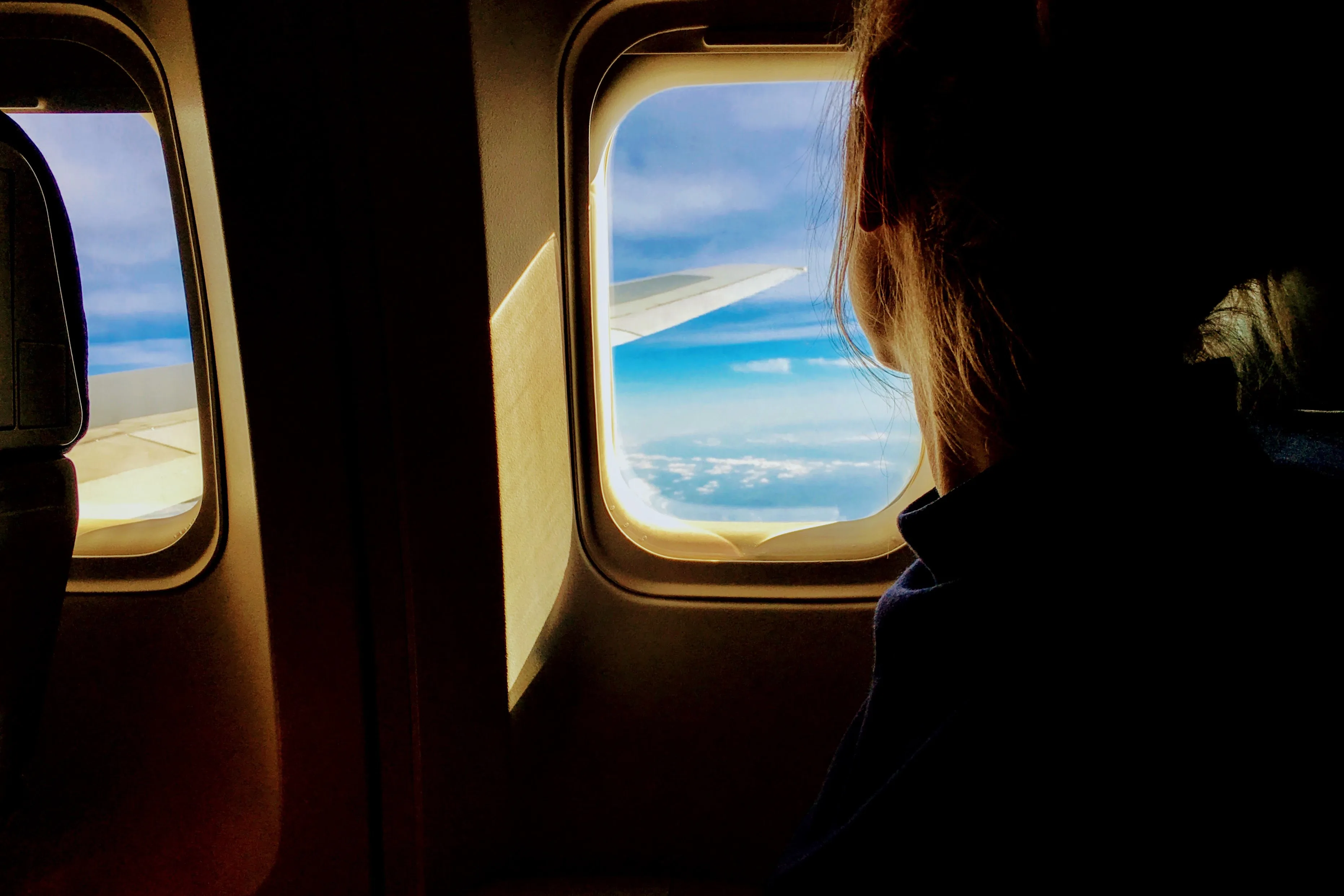 Vrouw kijkt uit het raam van een vliegtuig naar een blauwe lucht met wolken en de vleugel van het toestel.