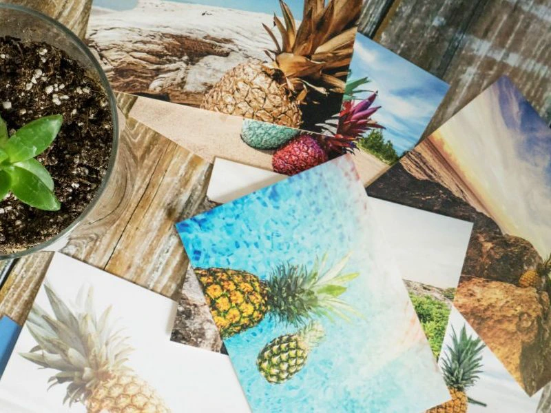 Close-up van een stapel ansichtkaarten met tropische afbeeldingen van ananassen en stranden, naast een pot met een kleine groene plant.