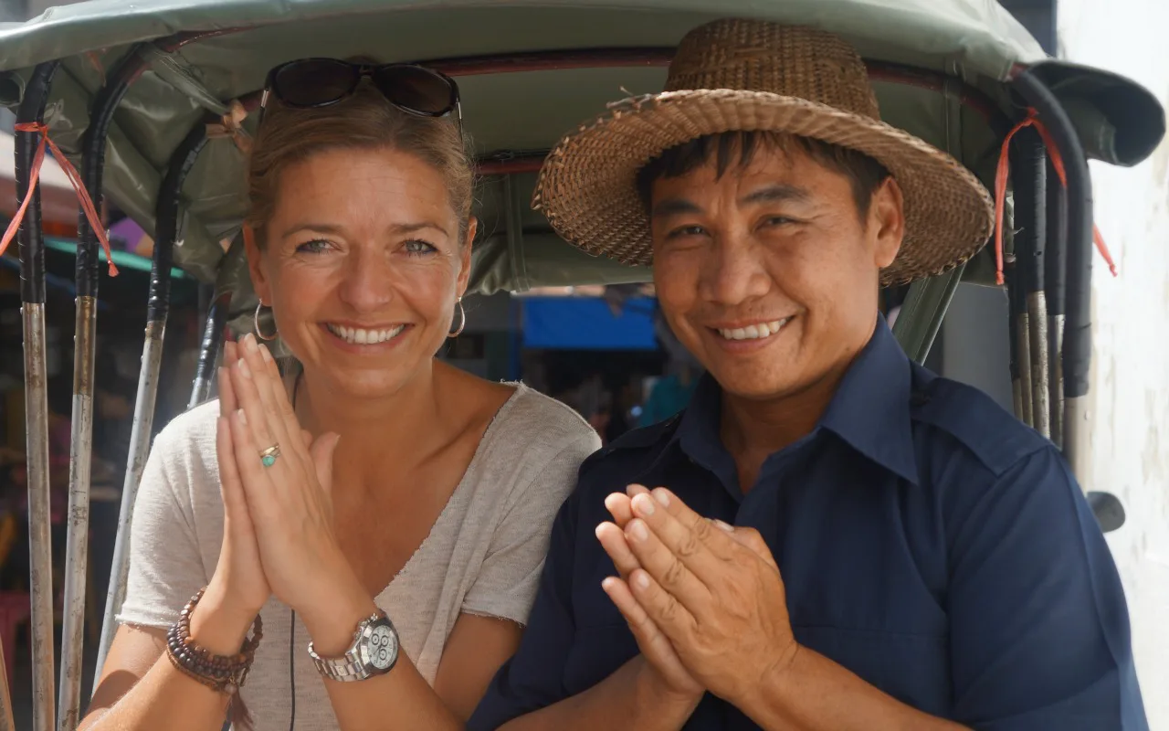 Vrouw en man glimlachen naar de camera en maken samen een begroetingsgebaar met de handen tegen elkaar.