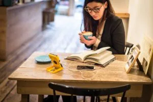 Vrouw met bril leest in een boek aan een houten tafel in een café, met een kop koffie in haar hand en een telefoon op een gele standaard.