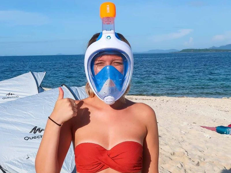 Vrouw op het strand met een rood bikini-topje en een volledig snorkelmasker, die lachend een duim omhoog steekt met de zee op de achtergrond.
