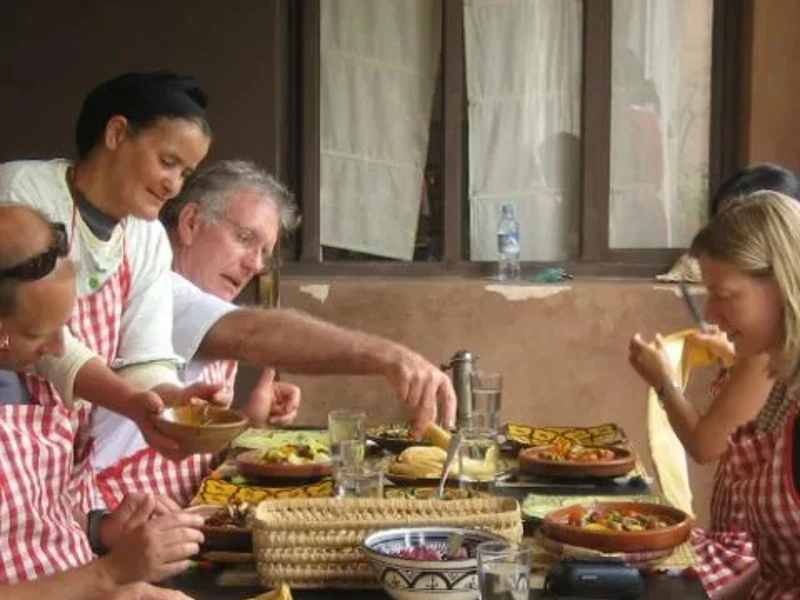 Groep reizigers en een lokale vrouw genieten samen van een maaltijd tijdens een kookworkshop, met kleurrijke gerechten op tafel en iedereen in rood-wit gestreepte schorten.