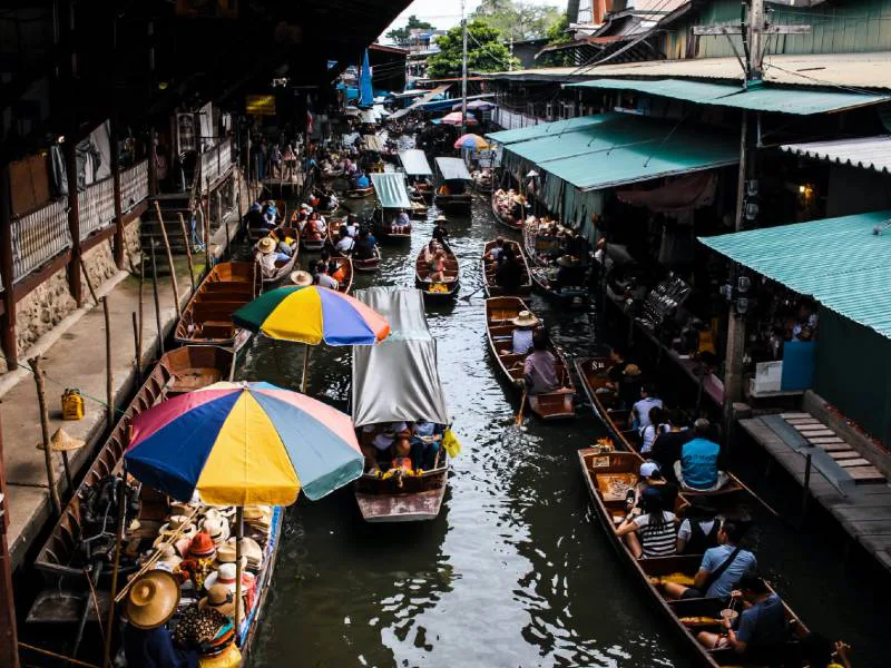 Drukke drijvende markt in Thailand met houten bootjes vol mensen en kleurrijke parasols, varend door een smalle waterweg tussen winkeltjes.